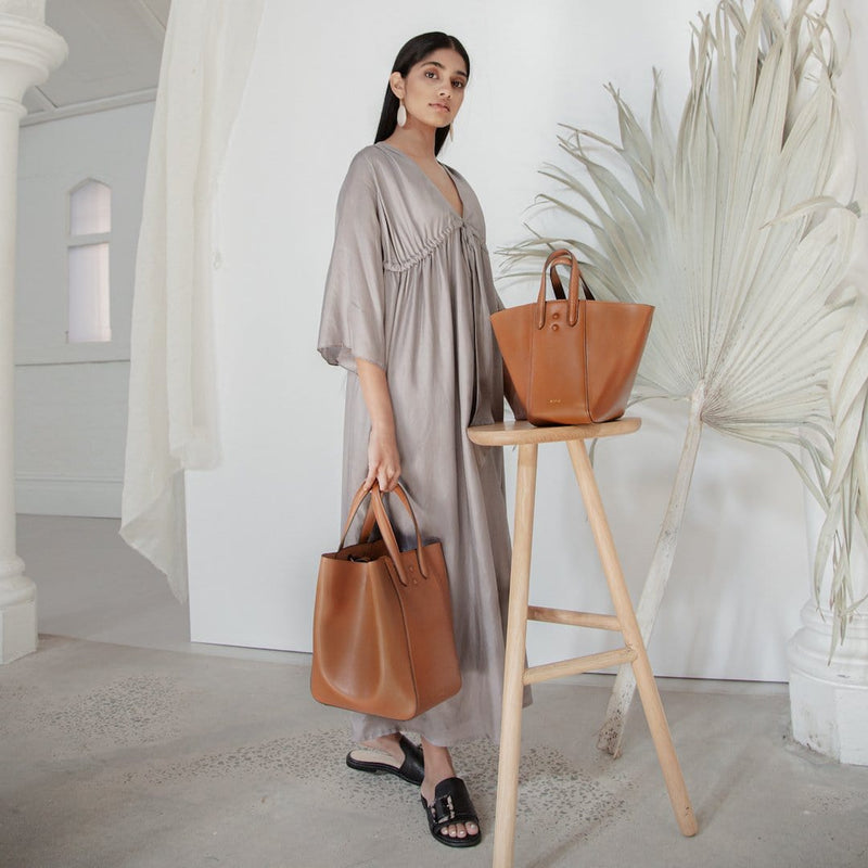 Woman standing next to stool shown with two luxury leather handbags, tan leather hybrid tote bucket bag, two different sizes, the regular Eight and Eight Mini.