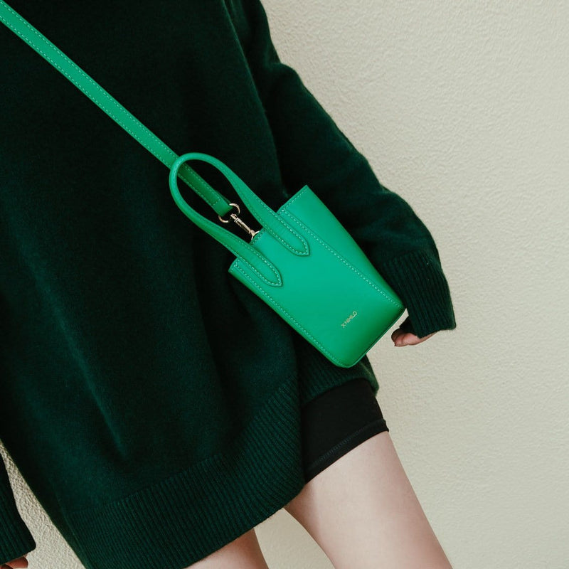 Woman in green sweater wearing Micro bright green leather bucket hybrid handbag crossbody, with detachable long strap, top handle and X NIHILO embossed in gold on the bottom front.