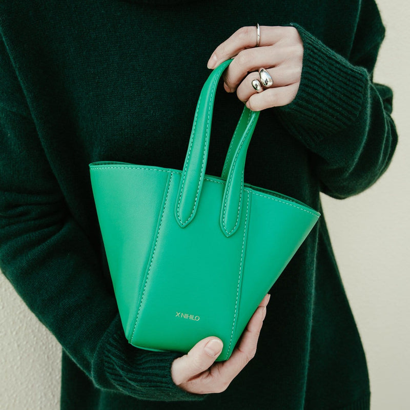 Woman in green sweater holding Micro bright green leather bucket hybrid handbag, with top handle and X NIHILO embossed in gold on the bottom front.