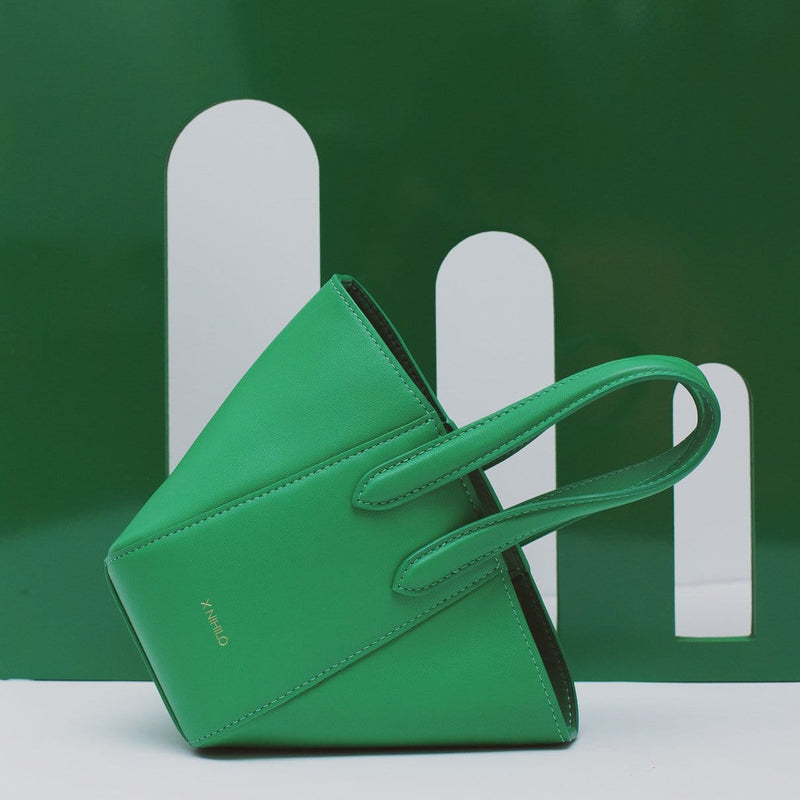 Micro bright green leather bucket hybrid handbag on its side, with top handle and X NIHILO embossed in gold on the bottom front shown in front of green backdrop with archways.