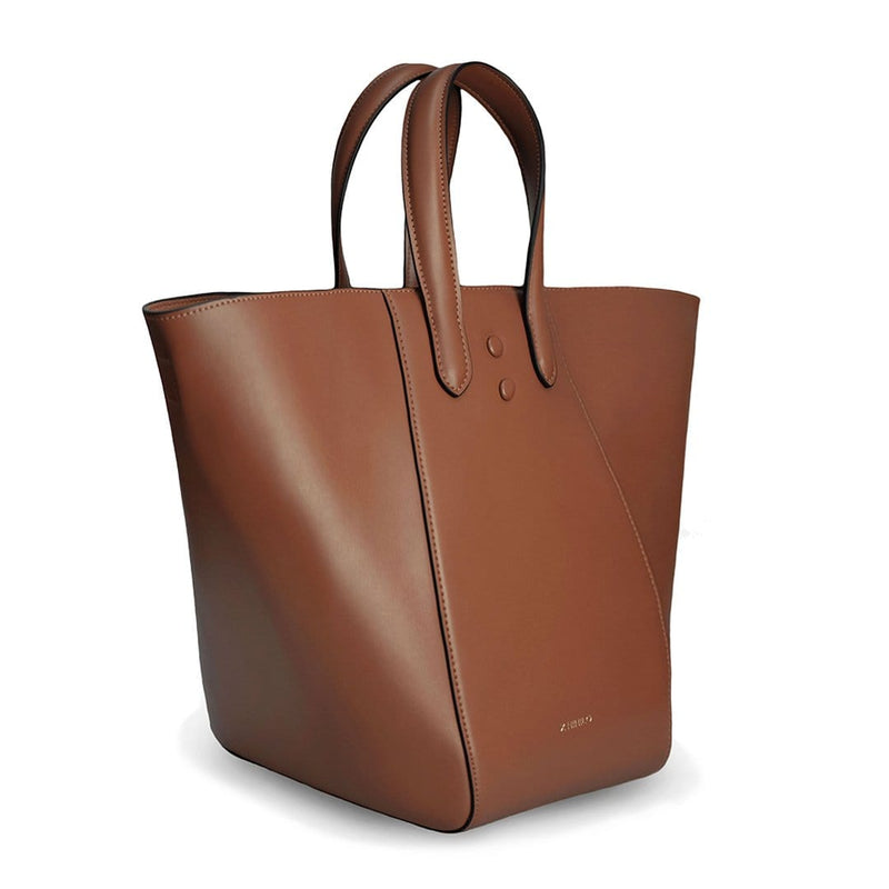 Angled side view of large luxury tan leather tote bucket bag, genuine nappa leather bag with handle and logo X NIIHILO embossed in gold on the bottom front, single button closure.