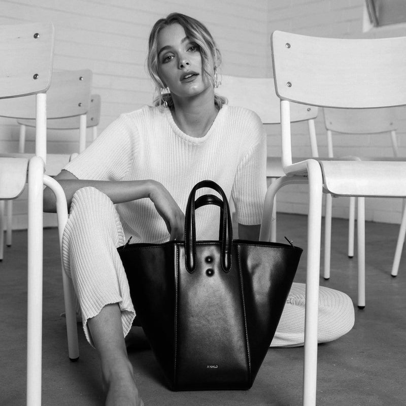 Woman sitting on the ground next to chairs holding a black genuine nappa leather handbags, luxury fashion bucket tote hybrid.