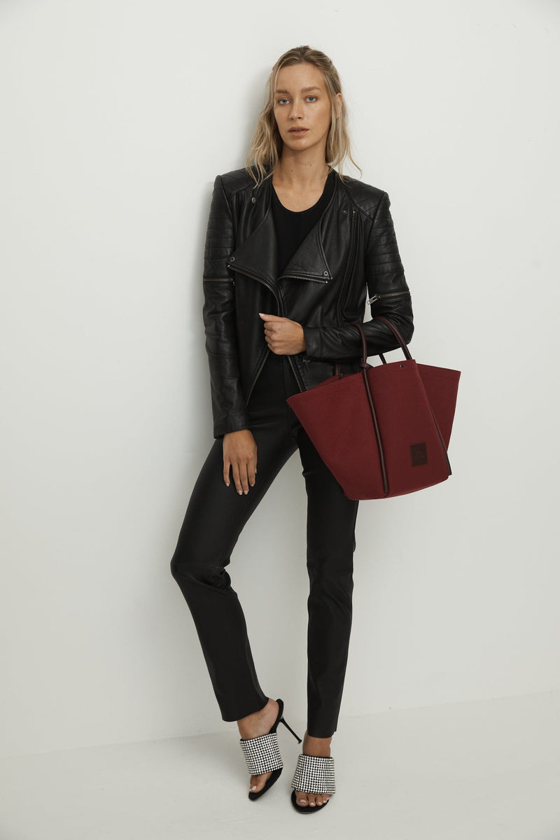 Woman holding a burgundy canvas tote bucket bag, with burgundy rolled leather handles, silver button closure at the top and small square logo of WEST14TH and X NIHILO. Luxury bags, made from hardwearing canvas and rolled leather.