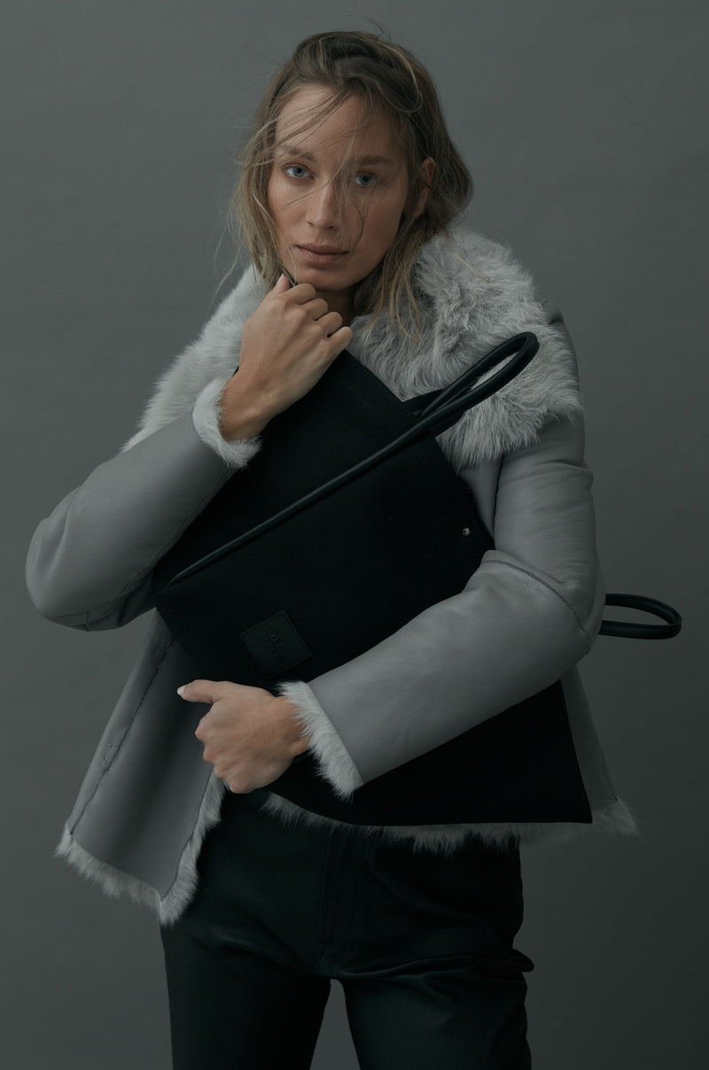 Women in grey jacket hugging a black canvas tote bucket bag with both of her arms, with black leather rolled leather handles, silver button closure at the top, silver hardware and small square logo of WEST14TH and X NIHILO.  Luxury bags, made from hardwearing canvas and rolled leather.