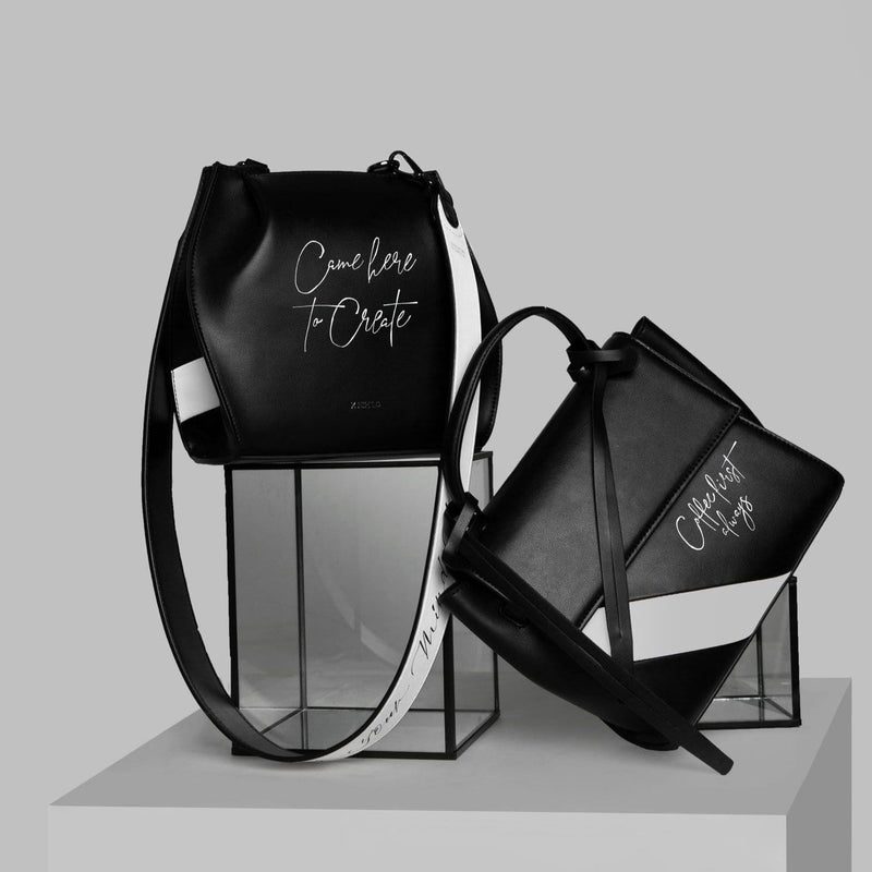 Hybrid mini bucket bag in black and white stripe leather, with gunmetal hardware, detachable and adjustable strap, with calligraphy "Came here to Create" written across the surface. Positioned next to a black and white stripe rectangle handbag leaning against a rectangular glass prism, with calligraphy "Coffee first always" written on it. 