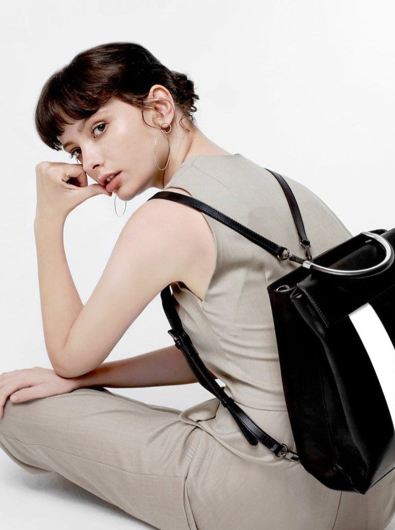 Woman sitting on the ground wearing X NIHILO Hunter in black and white stripe as a backpack bag, fashion bag with high shine gunmetal hardware, detachable and adjustable strap, two hidden magnetic clasps closure for front compartment, pockets fitted inside, and zip closure for back compartment, luxury cow nappa leather handbag, genuine leather bag