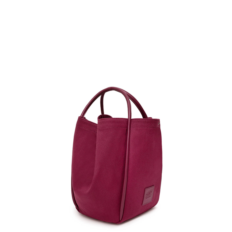 Side view of burgundy canvas tote bucket bag, with burgundy leather rolled leather handles, silver button closure at the top and small square logo of WEST14TH and X NIHILO. Shown as bucket bag, with button used to bring in wings.