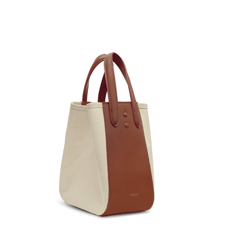 Side view of tan leather and natural canvas tote bucket bag, genuine nappa leather bag with handle and logo X NIIHILO embossed in gold on the bottom front.