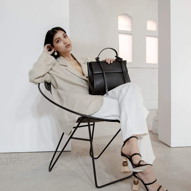 Full body shot of a woman leaning back in a chair holding rectangle genuine black leather handbag in a bright indoor setting.