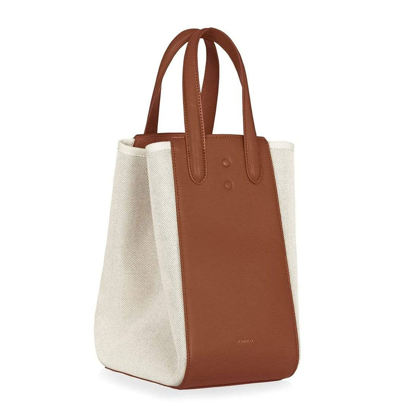 Angled side view of large tan leather and natural canvas fabric hybrid tote bucket bag, genuine nappa leather bag with handle and logo X NIIHILO embossed in gold on the bottom front, single button closure.