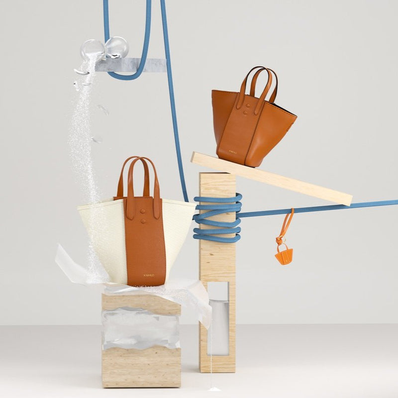 3D animation of two large genuine leather tote bucket bag hybrids, one in tan and the other in tan and canvas, shown in a balancing act on wood and stings.