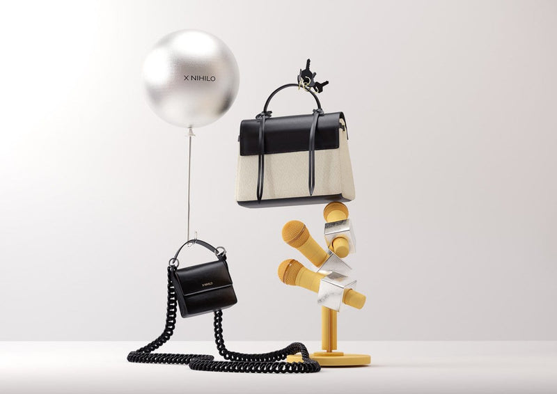 3D detailed artistic composition of floating black leather and canvas fabric trapezoid bag on top of three yellow abstract microphones and a mini black leather square bag with a black chain being held by a silver balloon with the X NIHILO name.