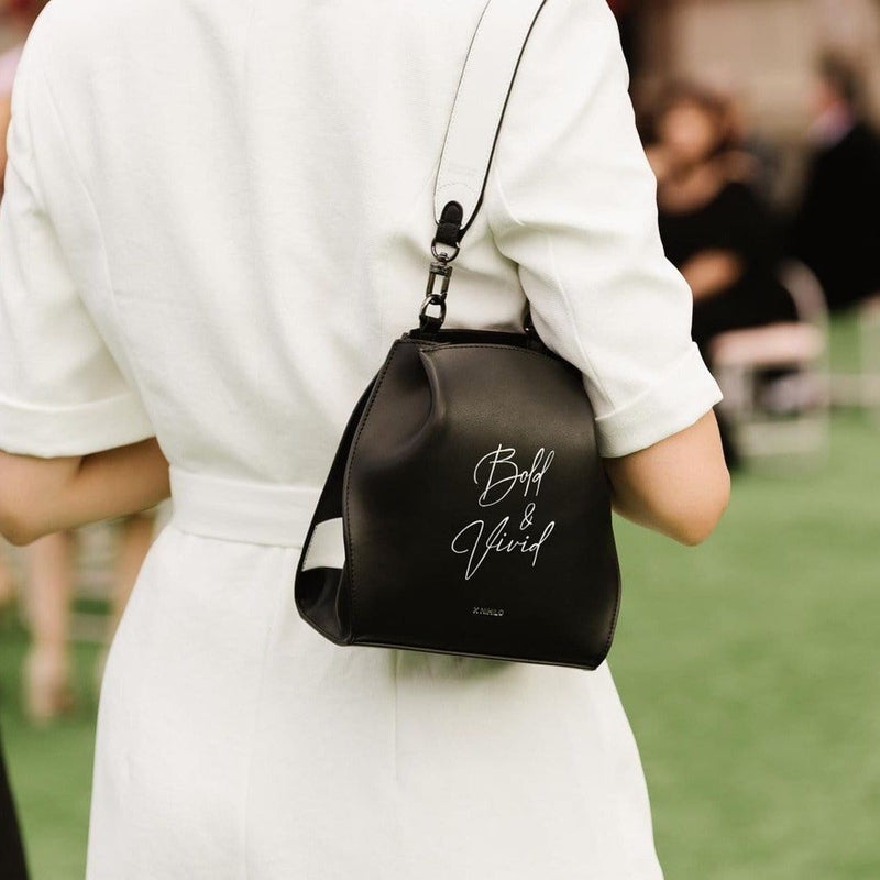 A woman with her back facing the camera wearing the black and white stripe leather hybrid mini bucket bag with a short white strap on one shoulder, "Bold & Vivid" written in calligraphy on it..