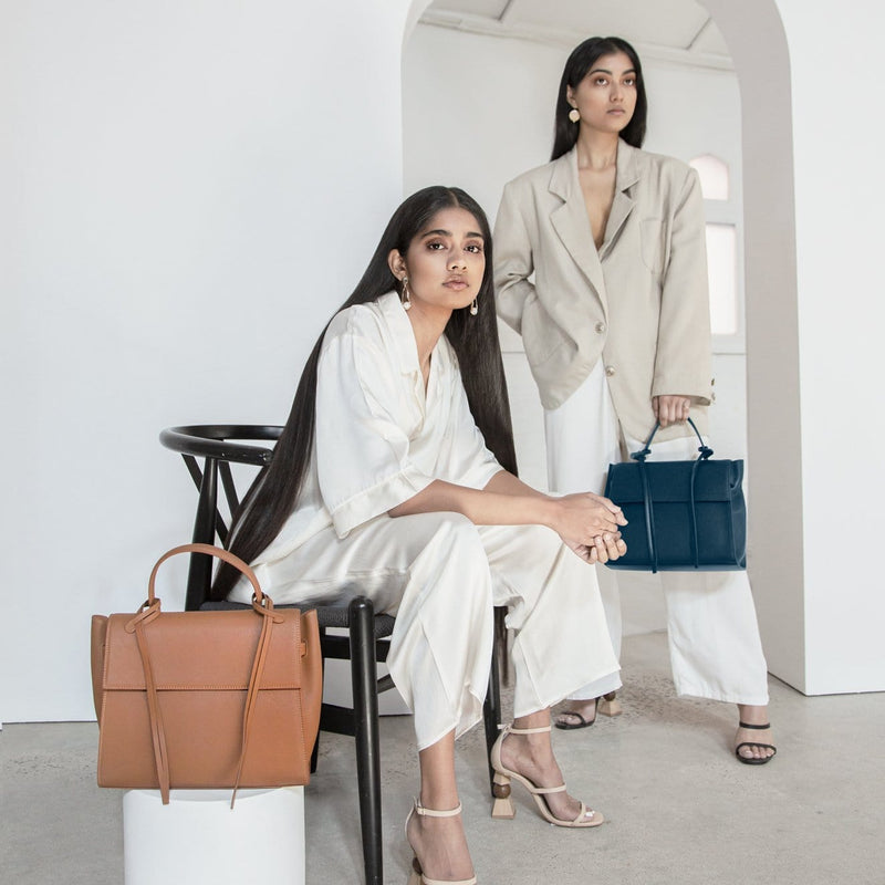 A woman wearing all white sitting in a chair, a rectangle genuine tan leather work bag and handbag with leather tassels, front flap and handle placed in the foreground. Another woman wearing a beige blazer standing in the background holding a rectangle navy blue genuine leather handbag with leather tassels, flap and handle. 