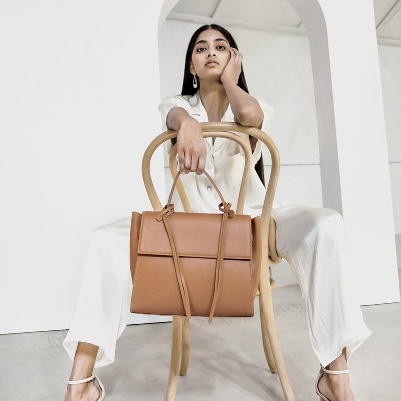 A woman wearing all white sitting in a chair, one hand on cheek, holding a rectangle genuine tan leather work bag and handbag with leather tassels, front flap and handle in the other hand to the front.