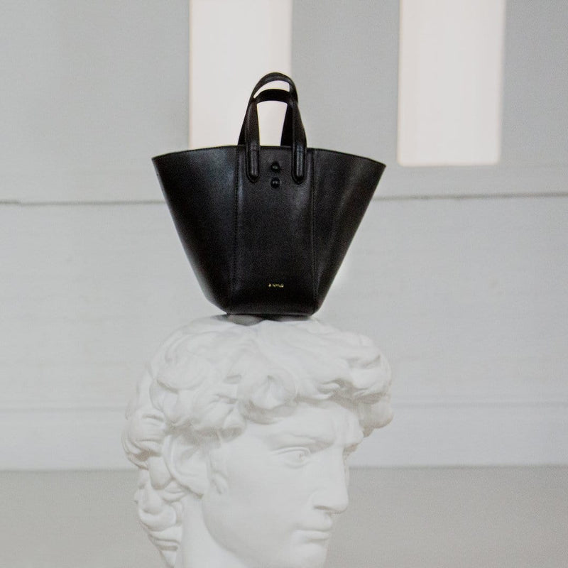 X NIHILO Eight mini in black bag, fashion bag with adjustable shoulder strap, single snap button top closure, and soft gold hardware, luxury cow nappa leather handbag, genuine leather bag shown balancing on white Roman sculpture of a head.