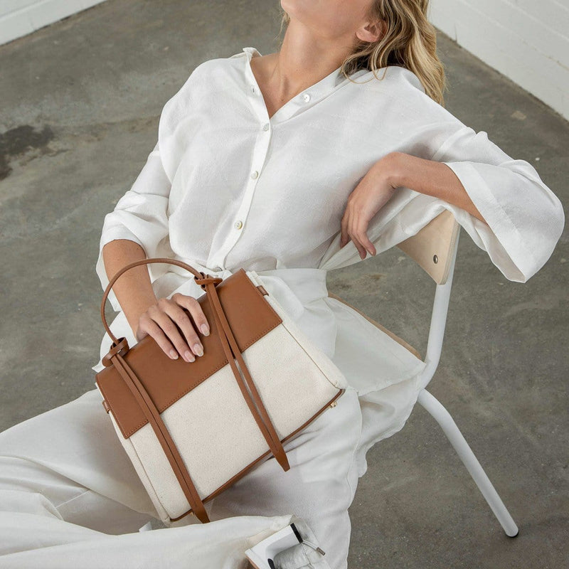 Woman wearing white blouse leaning backward on a chair, the image is cropped off from the head, she is holding a tan leather handbag with leather tassels, front flap and handle.