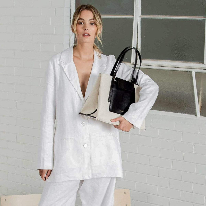 A woman wearing all white holding a rectangle black leather and natural canvas fabric fashion tote bag with black leather handle, standing in front of a white brick wall.