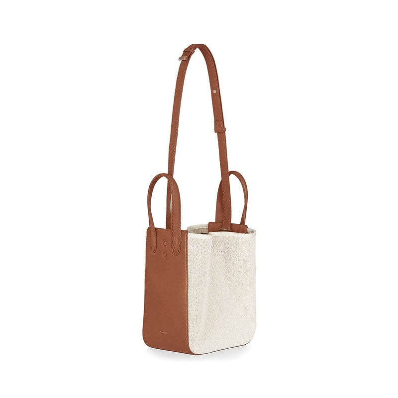 Angled side view of large tan leather and natural canvas fabric hybrid tote bucket bag, genuine nappa leather bag with handle and logo X NIIHILO embossed in gold on the bottom front, single button closure with detachable strap at full length.