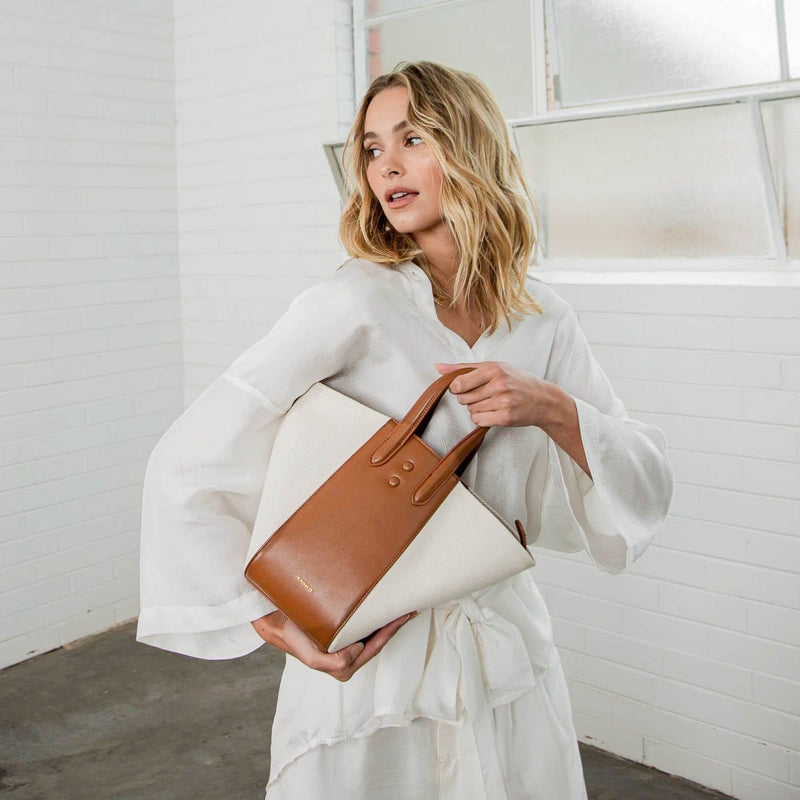 Woman wearing white holding Mini tan leather and natural canvas tote bucket bag, genuine nappa leather bag with handle and logo X NIIHILO embossed in gold on the bottom front.
