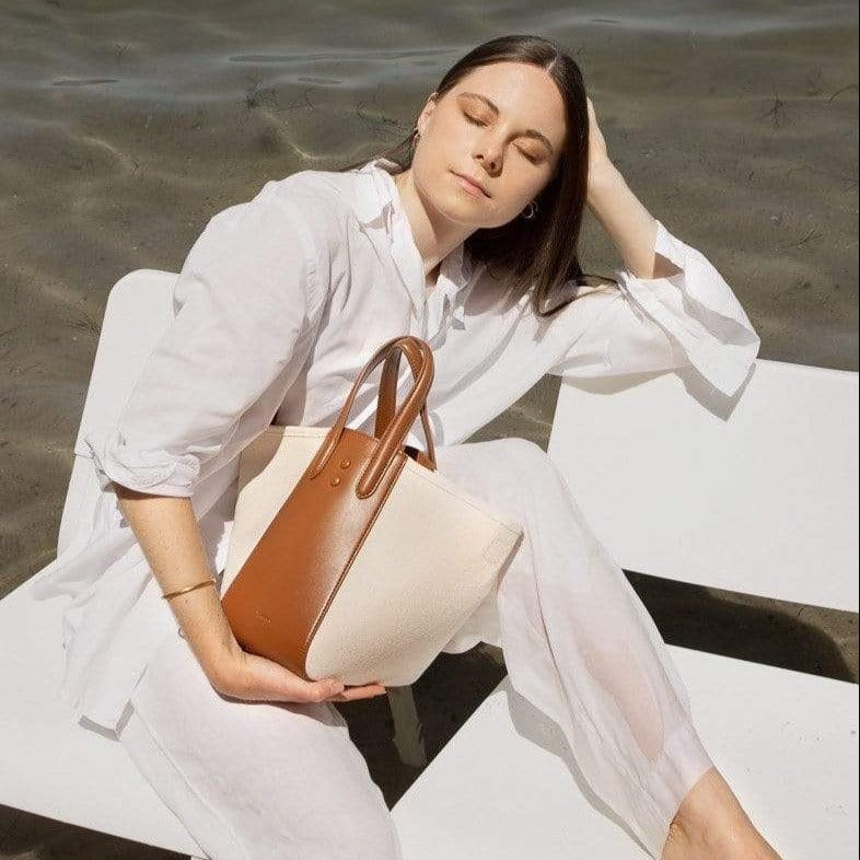 Woman by the ocean wearing white holding Mini tan leather and natural canvas tote bucket bag, genuine nappa leather bag with handle and logo X NIIHILO embossed in gold on the bottom front.