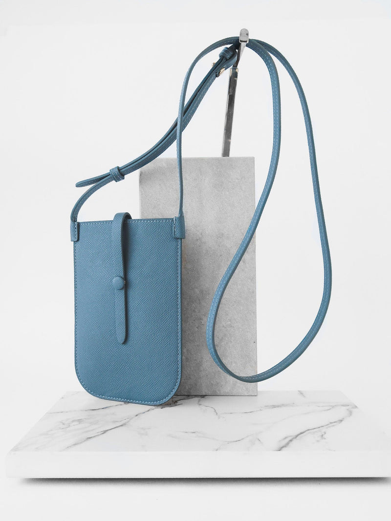 Rectangular Chambray blue phone pouch with opening on top and a attached shoulder strap against white marmol stand.