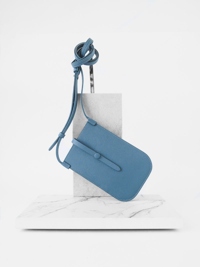 Rectangular Chambray blue phone pouch with opening on top and a attached shoulder strap against white marmol stand