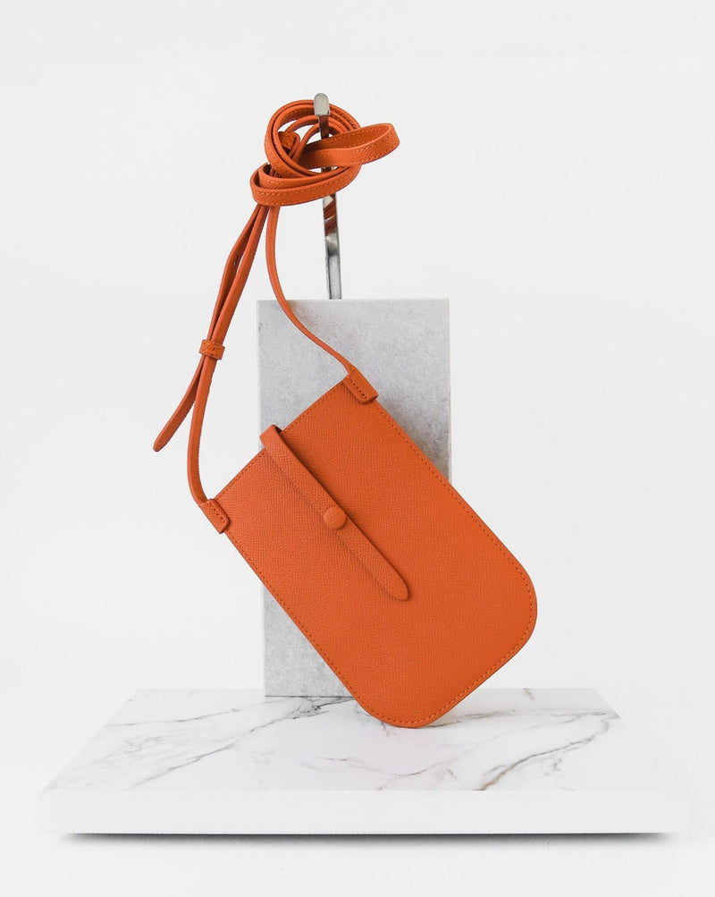 Rectangular Tangerine orange phone pouch with opening on top and an attached shoulder strap. Standing against a white marmol stand.