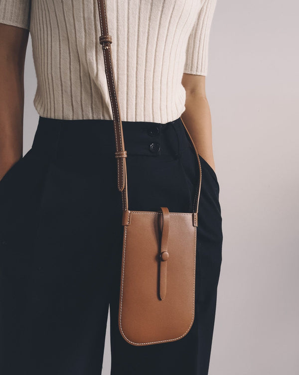 Model wearing rectangular woman's tan phone pouch. White stitching along the edges and an opening for the phone at the top. A sling with a tan button keeps the essentials secured.