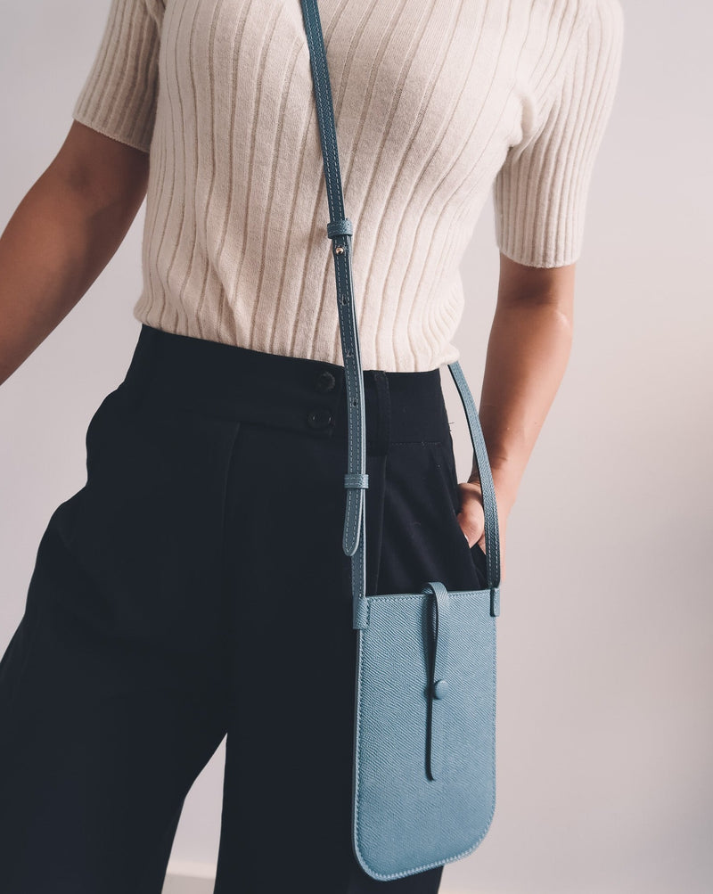 Model wearing rectangular Chambray blue phone pouch with opening on top and an attached shoulder strap.