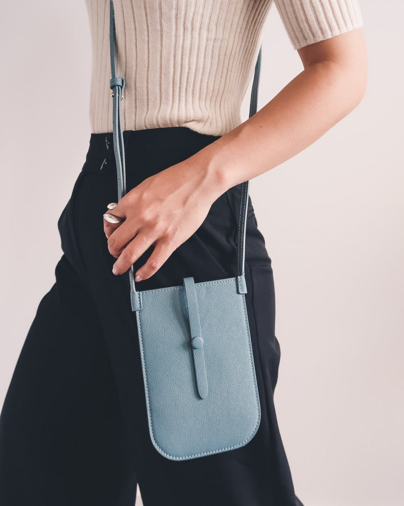 Model wearing rectangular Chambray blue phone pouch with opening on top and a attached shoulder strap.