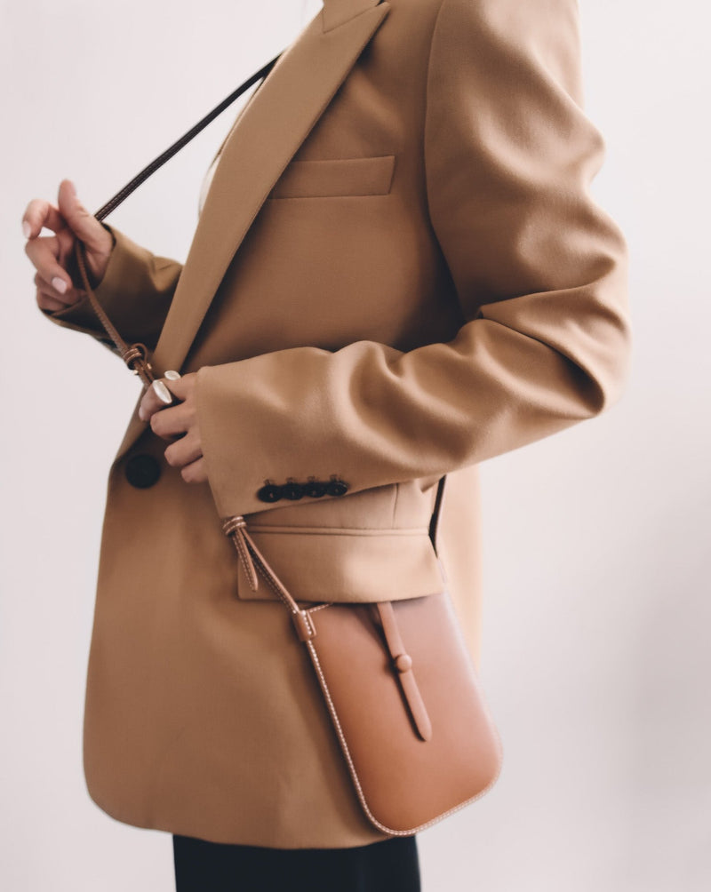 Model wearing rectangular woman's tan phone pouch. White stitching along the edges and an opening for the phone at the top. A sling with a tan button keeps the essentials secured.