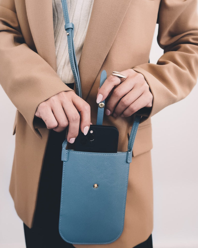 Model wearing rectangular Chambray blue phone pouch with opening on top and an attached shoulder strap.