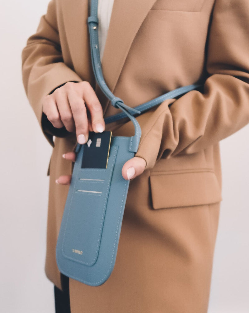 Model wearing rectangular Chambray blue phone pouch with opening on top and a attached shoulder strap. Back view, card sliding in card holder.