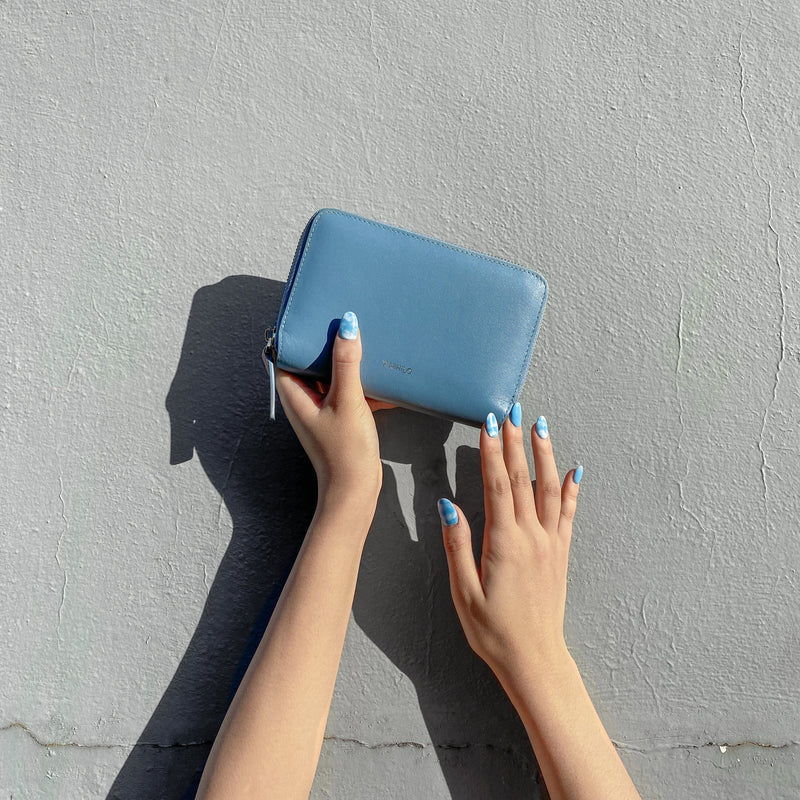 Two hands of a female with light-blue nail polish, one hand is holding the sky blue leather passport wallet against a white wall.