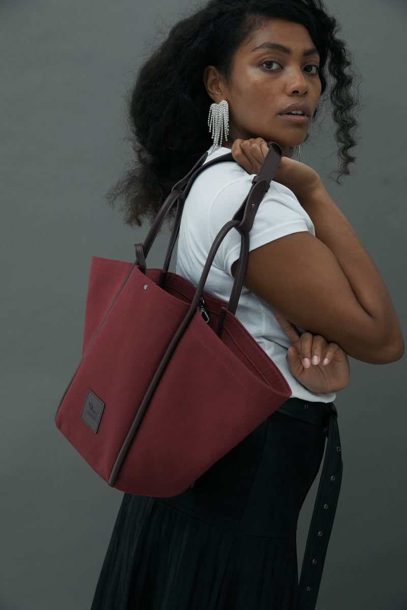 Close up shot of woman holding a burgundy canvas tote bucket bag, with burgundy rolled leather handles, silver button closure at the top and small square logo of WEST14TH and X NIHILO. Luxury bags, made from hardwearing canvas and rolled leather.