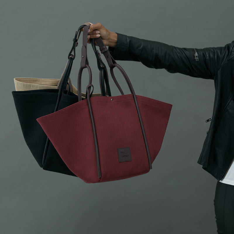 Woman's arm held out, holding three canvas tote bucket bags in camel, black and burgundy. Each with different coloured rolled leather handles , silver button closure at the top and small square logo of WEST14TH and X NIHILO at the front bottom.Luxury bags, made from hardwearing canvas and rolled leather.