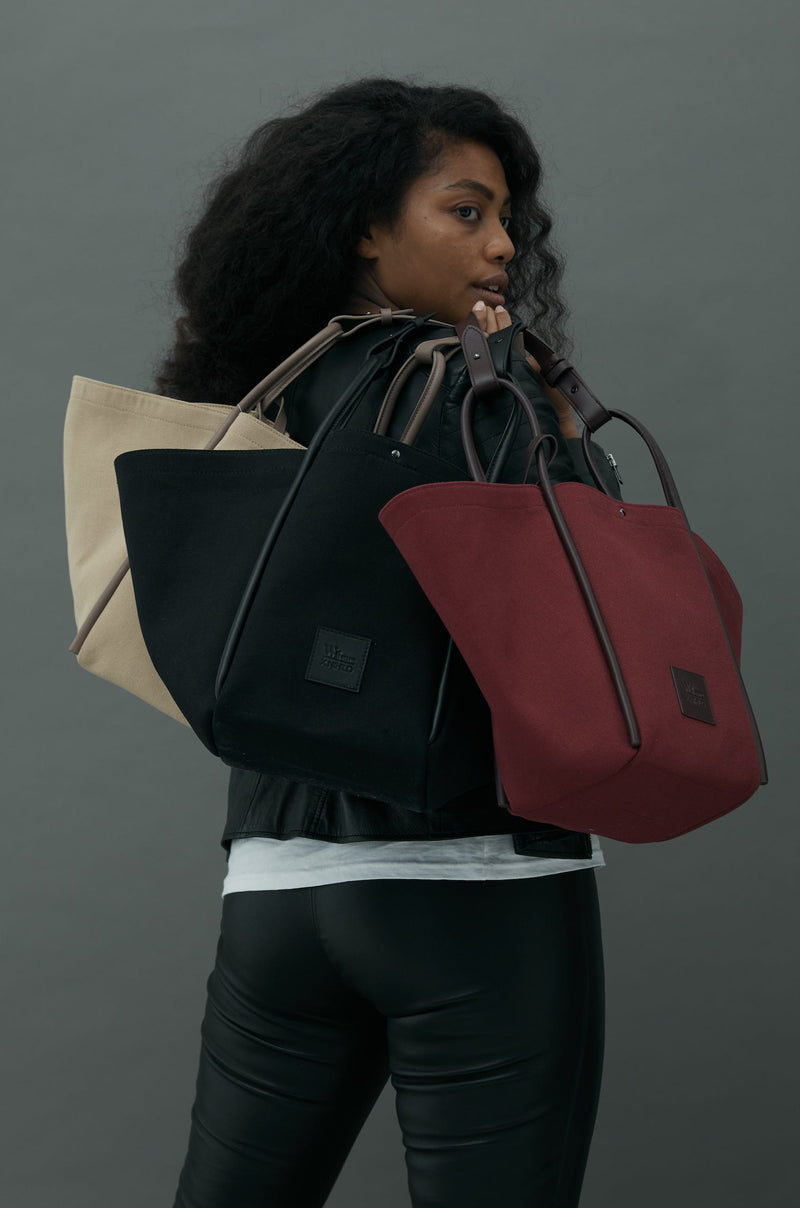  Woman turning her head towards the camera, holding three canvas tote bucket bags in camel, black and burgundy. Each with different coloured rolled leather handles , silver button closure at the top and small square logo of WEST14TH and X NIHILO at the front bottom.Luxury bags, made from hardwearing canvas and rolled leather.