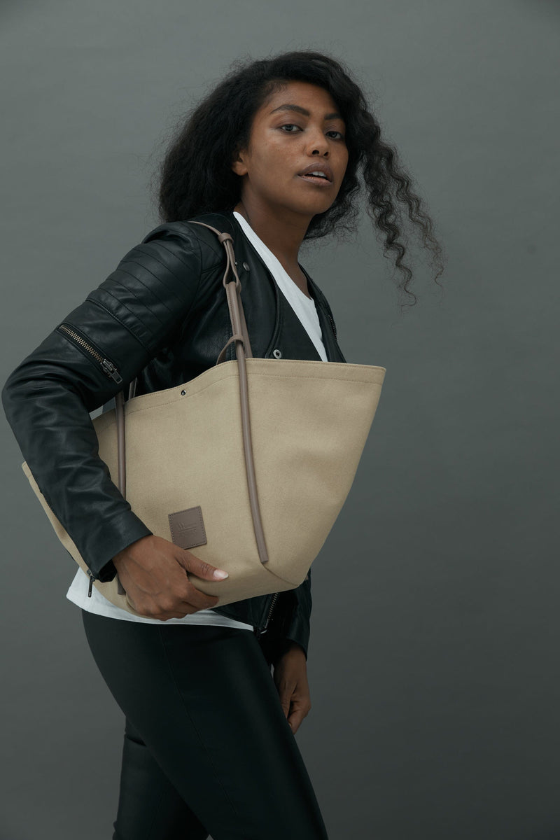 Woman wearing a Camel canvas tote bucket bag over her shoulder, with camel rolled leather handles, silver button closure at the top and small square logo of WEST14TH and X NIHILO. Luxury bags, made from hardwearing canvas and rolled leather.