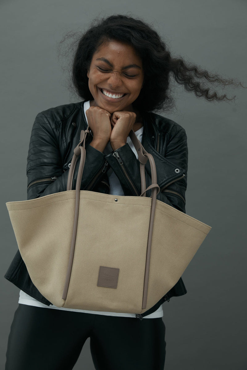 Woman smiling, holding a Camel canvas tote bucket bag, with camel rolled leather handles, silver button closure at the top and small square logo of WEST14TH and X NIHILO. Luxury bags, made from hardwearing canvas and rolled leather.