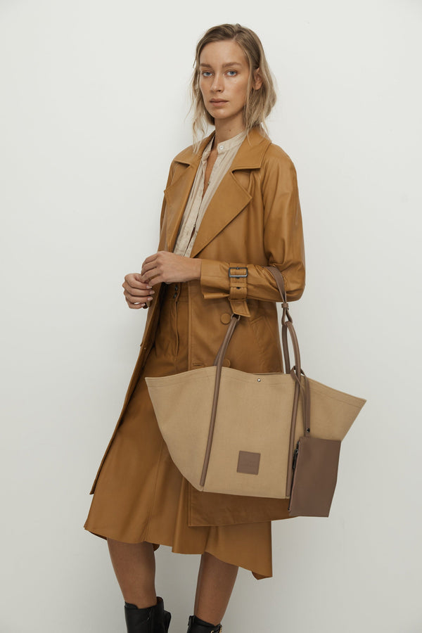 Woman holding a Camel canvas tote bucket bag, with camel rolled leather handles, silver button closure at the top and small square logo of WEST14TH and X NIHILO. Luxury bags, made from hardwearing canvas and rolled leather.
