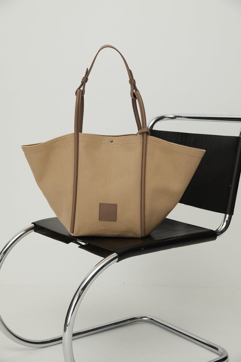 Camel canvas tote bucket bag sitting on a black chair, with camel rolled leather handles, silver button closure at the top and small square logo of WEST14TH and X NIHILO