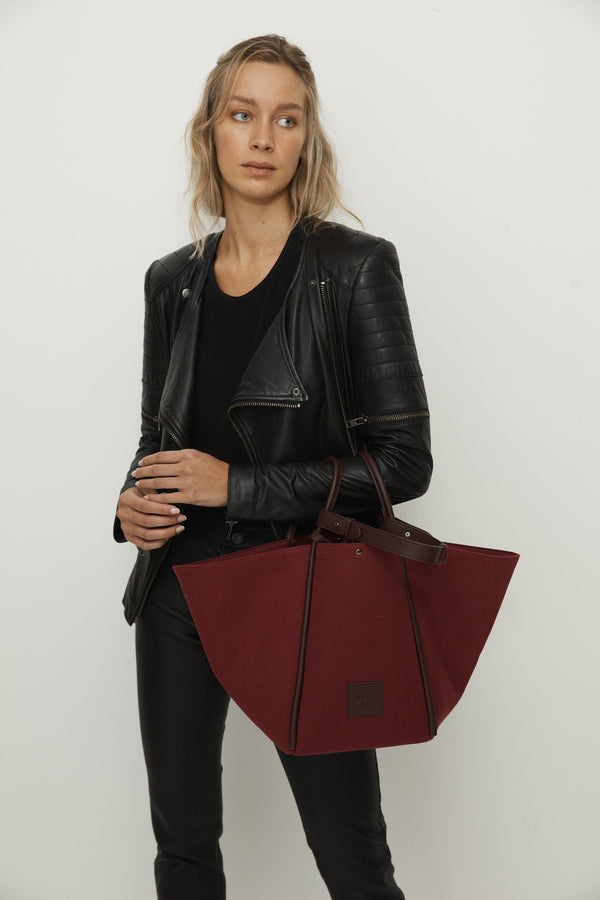 Woman holding a burgundy canvas tote bucket bag, with burgundy rolled leather handles, silver button closure at the top and small square logo of WEST14TH and X NIHILO. Luxury bags, made from hardwearing canvas and rolled leather