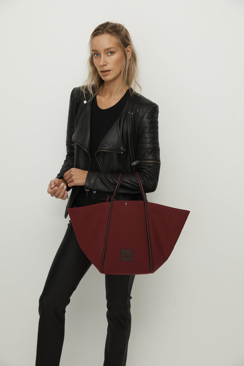 Woman holding a burgundy canvas tote bucket bag, with burgundy rolled leather handles, silver button closure at the top and small square logo of WEST14TH and X NIHILO. Luxury bags, made from hardwearing canvas and rolled leather.