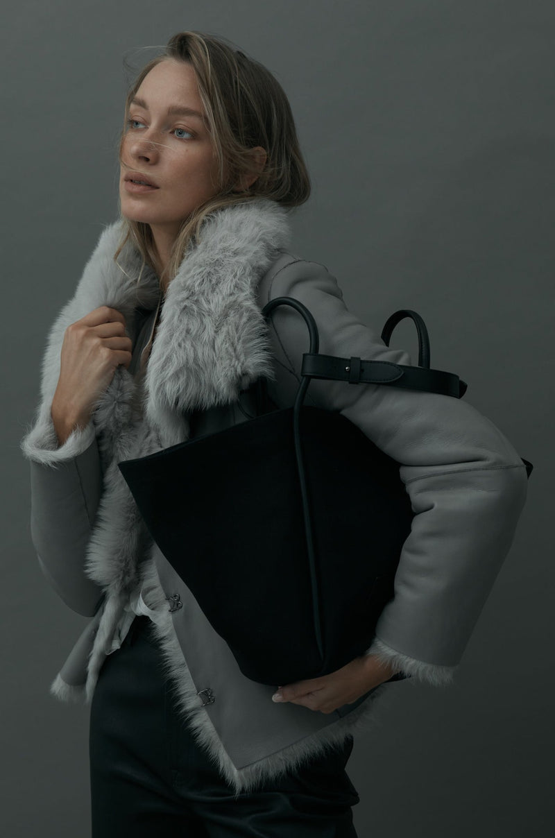 Women in grey jacket holding a black canvas tote bucket bag under her arm, with black leather rolled leather handles, silver button closure at the top, silver hardware and small square logo of WEST14TH and X NIHILO.