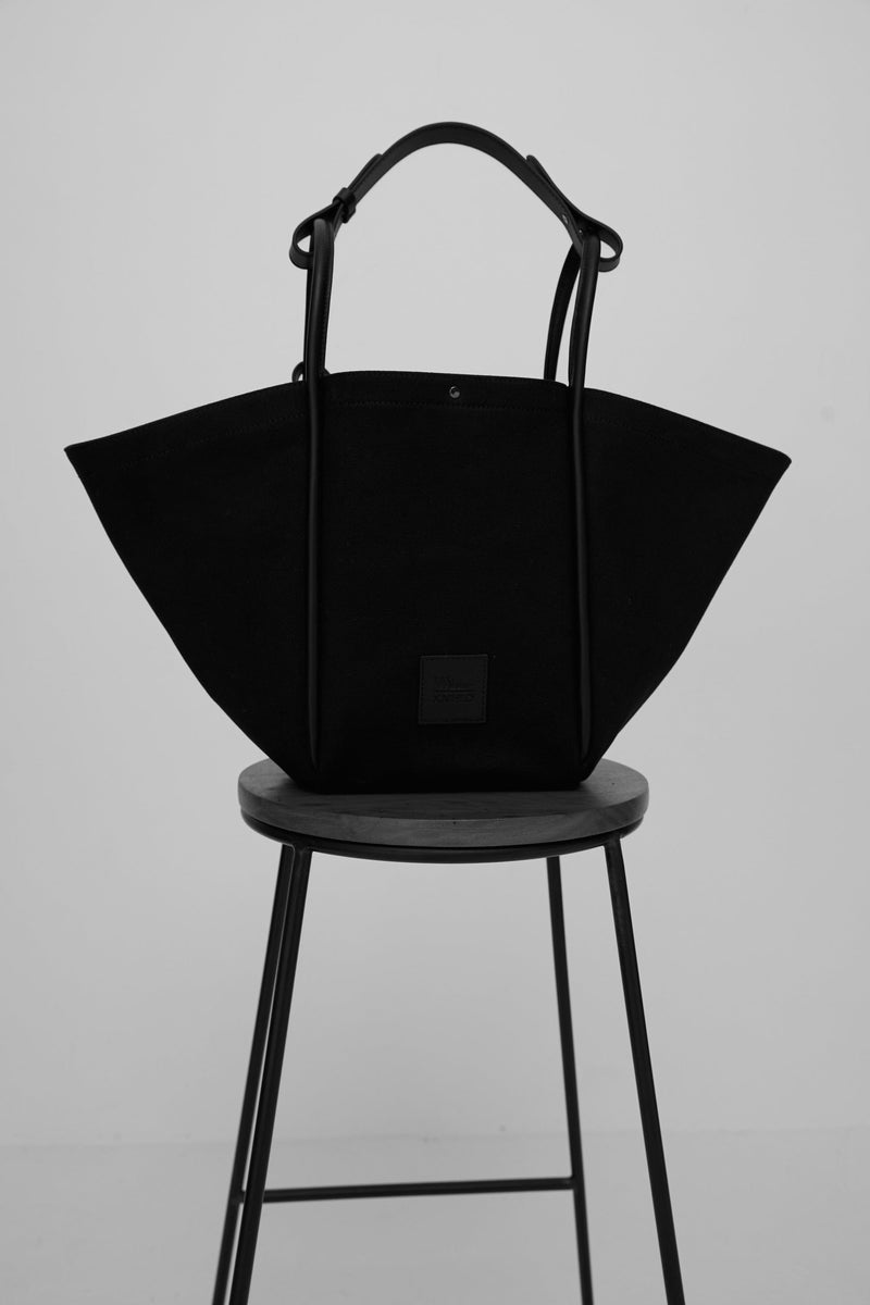 Black canvas tote bucket bag sitting on a stool, with black rolled leather handles, silver button closure at the top and small square logo of WEST14TH and X NIHILO.