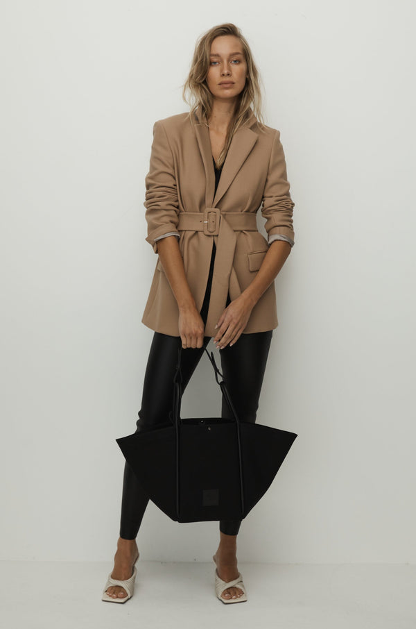 Women in camel jacket holding a black canvas tote bucket bag, with black leather rolled leather handles, silver button closure at the top and small square logo of WEST14TH and X NIHILO. 