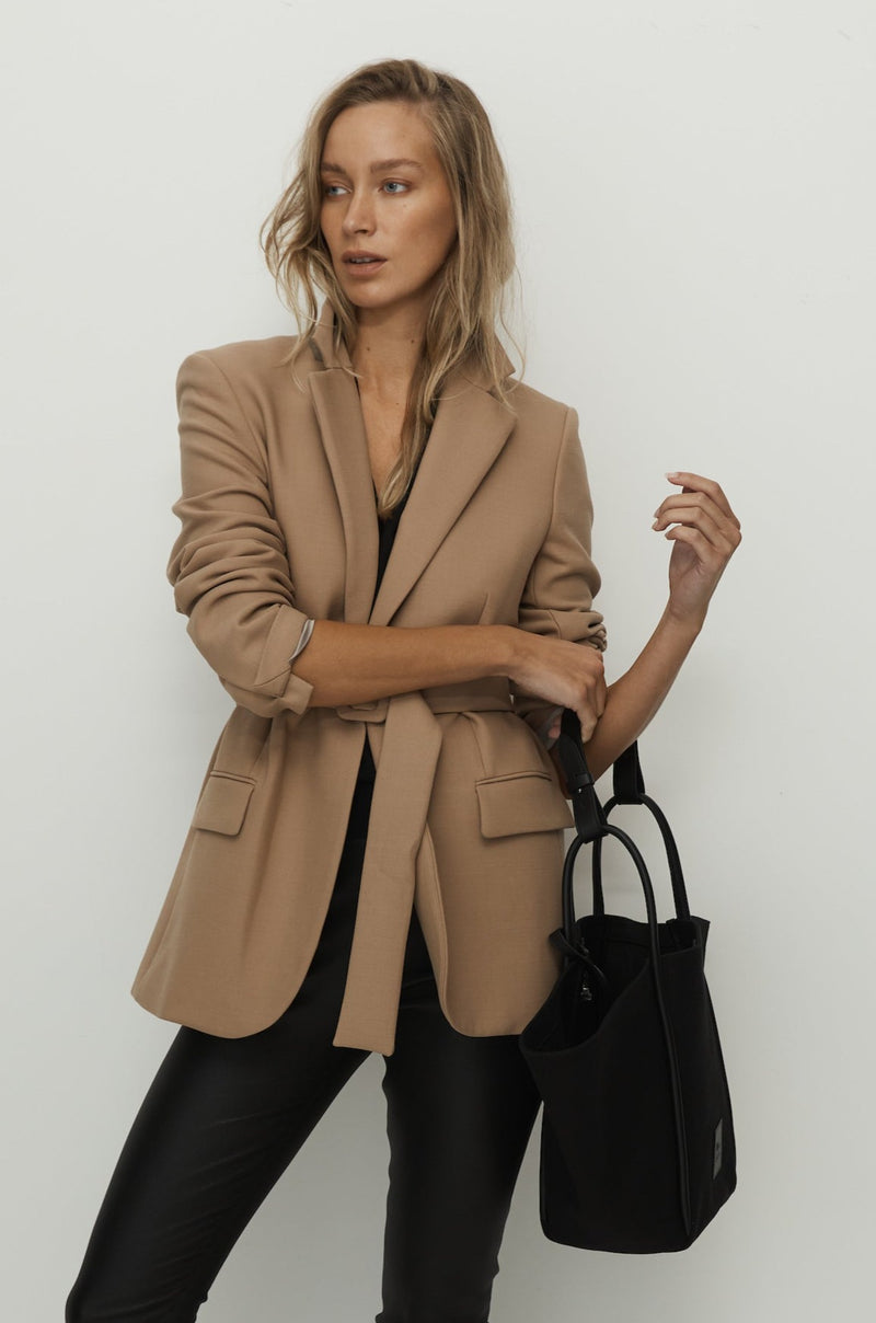 Women in camel jacket holding a black canvas tote bucket bag in the crook of her elbow, with black rolled leather handles, silver button closure at the top and small square logo of WEST14TH and X NIHILO. Luxury bags, made from hardwearing canvas and rolled leather.