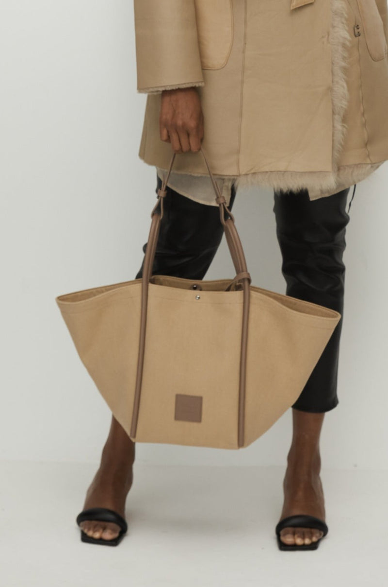Woman holding a Camel canvas tote bucket bag, with camel rolled leather handles, silver button closure at the top and small square logo of WEST14TH and X NIHILO. Luxury bags, made from hardwearing canvas and rolled leather.