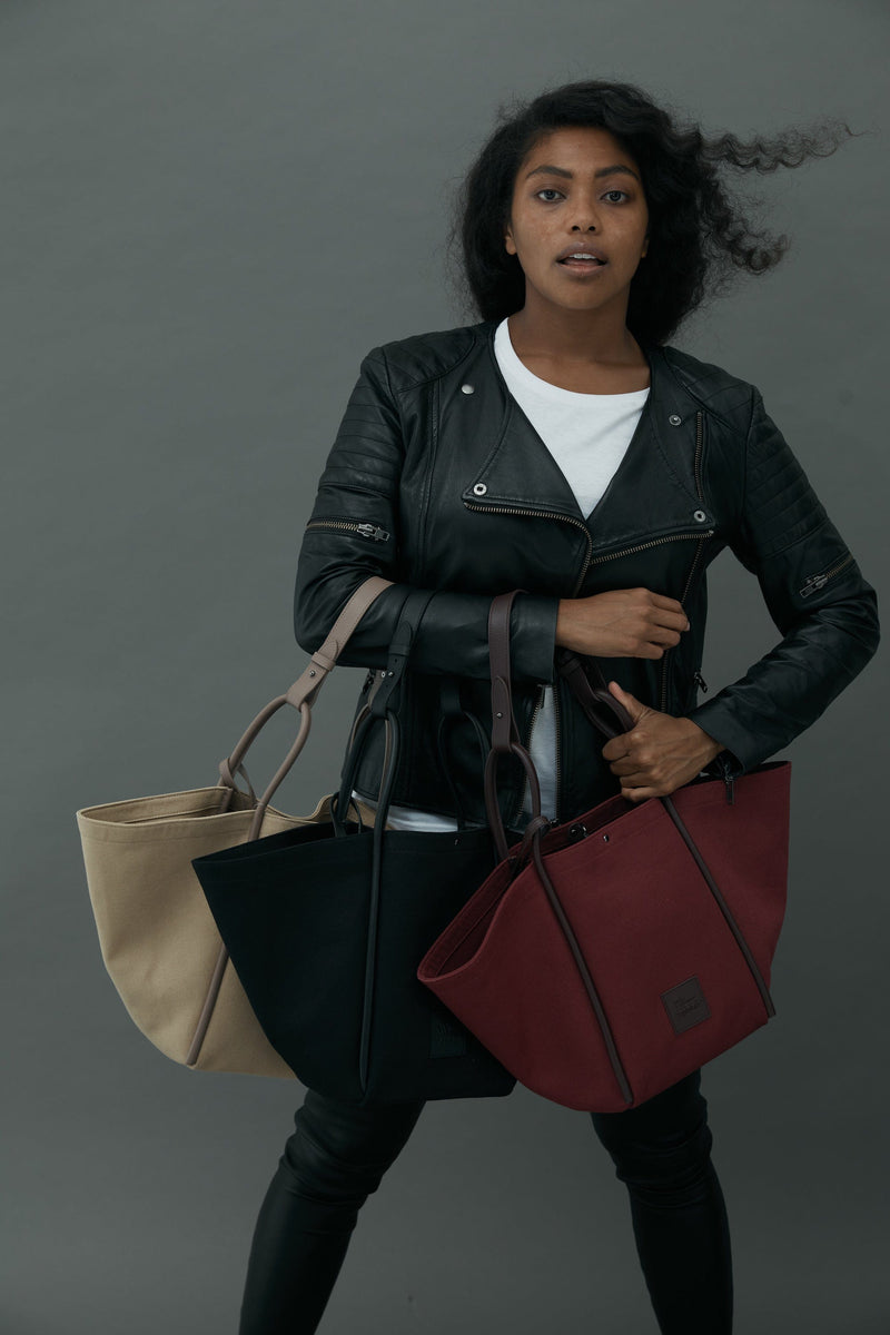 Woman holding three canvas tote bucket bags in camel, black and burgundy. Each with different coloured rolled leather handles , silver button closure at the top and small square logo of WEST14TH and X NIHILO at the front bottom.Luxury bags, made from hardwearing canvas and rolled leather.